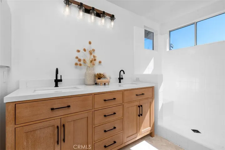 Primary Bathroom: Double-sink vanity with warm wood cabinetry, black fixtures, and a bright white countertop sits beside a tiled walk-in shower with natural light from clerestory windows.
