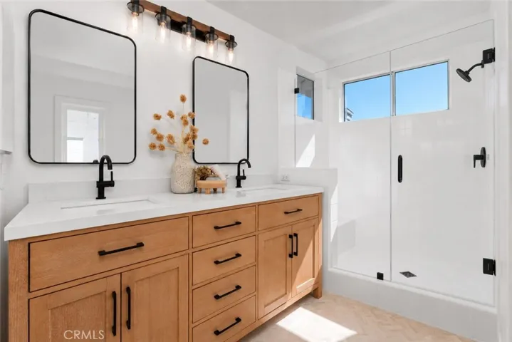 Virtually Enhanced - Primary Bathroom: Double-sink vanity with warm wood cabinetry, black fixtures, and a bright white countertop sits beside a tiled walk-in shower with natural light from clerestory windows.