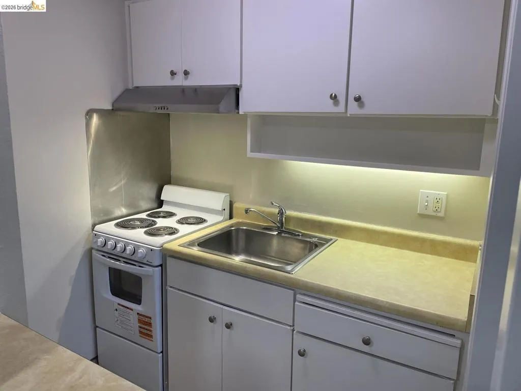 Compact kitchen featuring white cabinetry, a stainless steel sink, a four-burner electric range with an oven, and a range hood