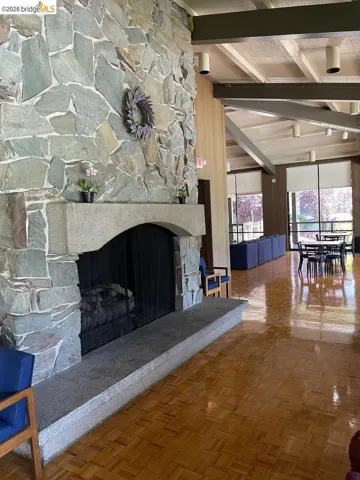 Stone-clad fireplace with a hearth, featuring wood-finish flooring, exposed beam ceiling, and large windows with roller shades
