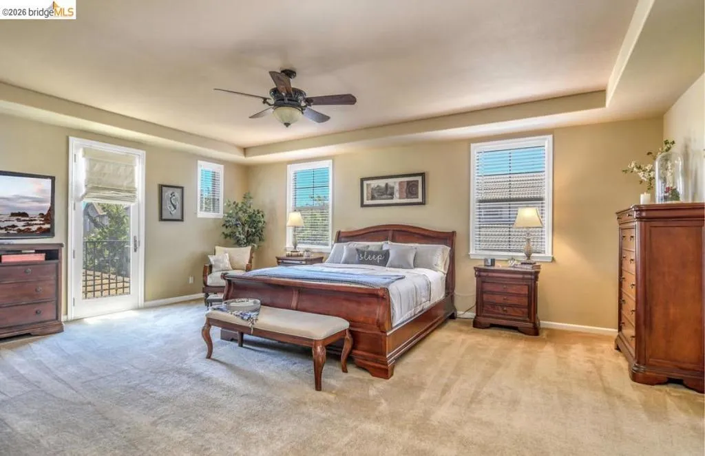 Spacious room featuring tray ceilings, carpeted flooring, multiple windows with blinds, a ceiling fan, and a glass-paneled door opening to a balcony