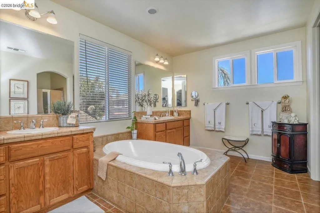Bathroom featuring an oval drop-in tub with tile surround, dual wood-finish vanities with tile countertops, and ceramic tile flooring