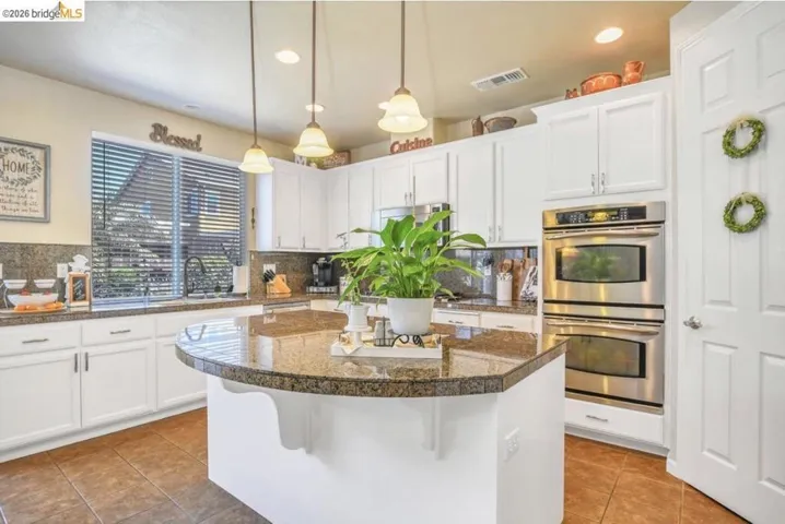 Kitchen featuring white cabinetry with brushed nickel hardware, a multi-toned granite countertop island with pendant lighting, a stacked stainless steel double oven, and a double-basin undermount sink with a gooseneck faucet