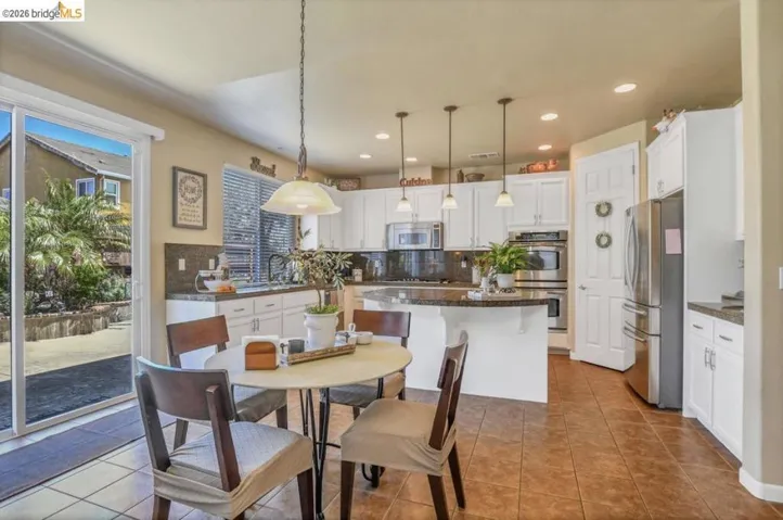 Open-concept kitchen featuring white cabinetry, dark stone countertops, stainless steel appliances, a center island with pendant lighting, and tile flooring