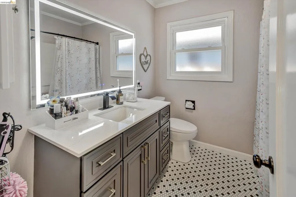 Bathroom featuring a grey vanity with a white countertop, an illuminated mirror, a white toilet, and black and white hexagonal floor tiling
