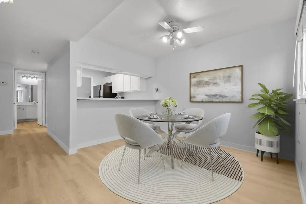Open-concept dining and kitchen area featuring a round glass-top table with modern seating, wood-finish flooring throughout, and a streamlined white cabinetry kitchen with stainless steel appliances
