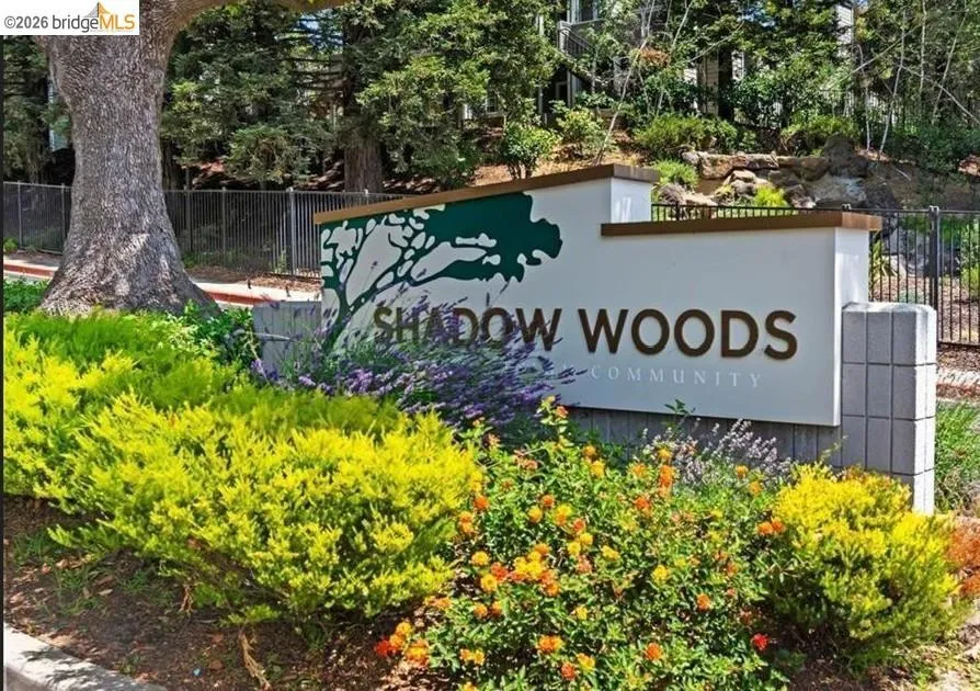 Community entrance sign featuring a decorative tree silhouette, block stone pillar, and surrounding shrubbery with colorful blooms