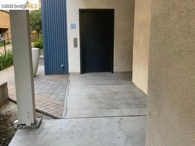 Concrete walkway leading to an entry featuring textured stucco walls, a dark panel door, and blue vertical siding
