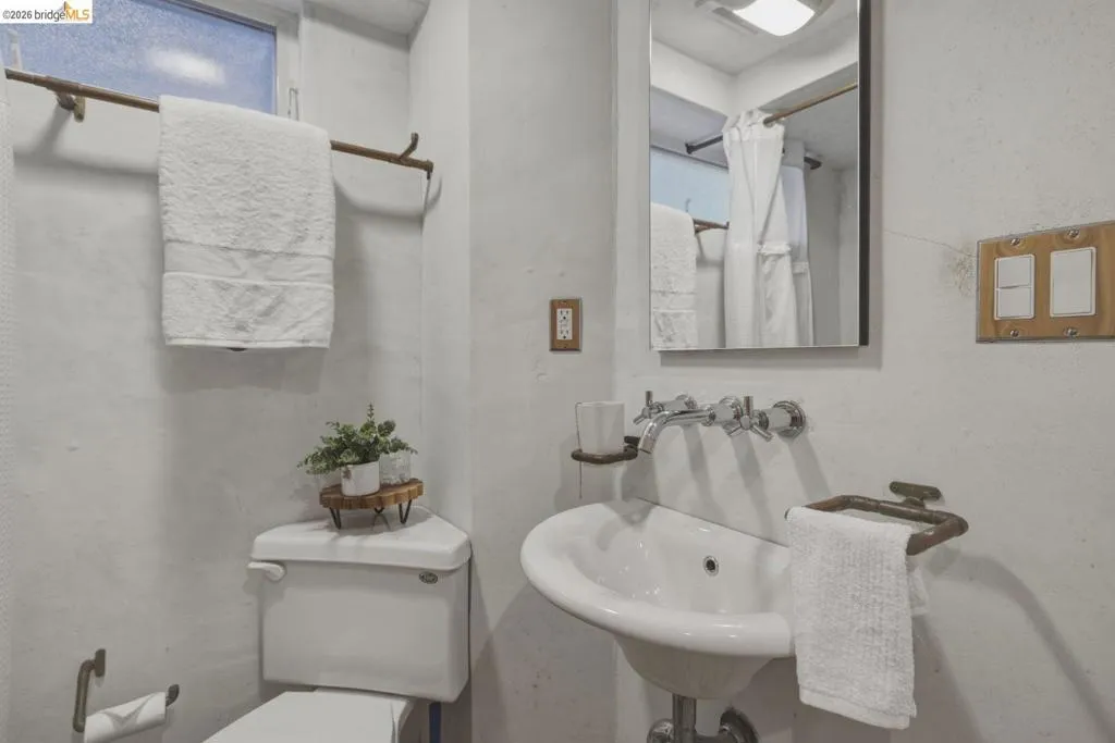 Compact bathroom featuring a wall-mounted sink with dual chrome faucets, a toilet, and a window with frosted glass