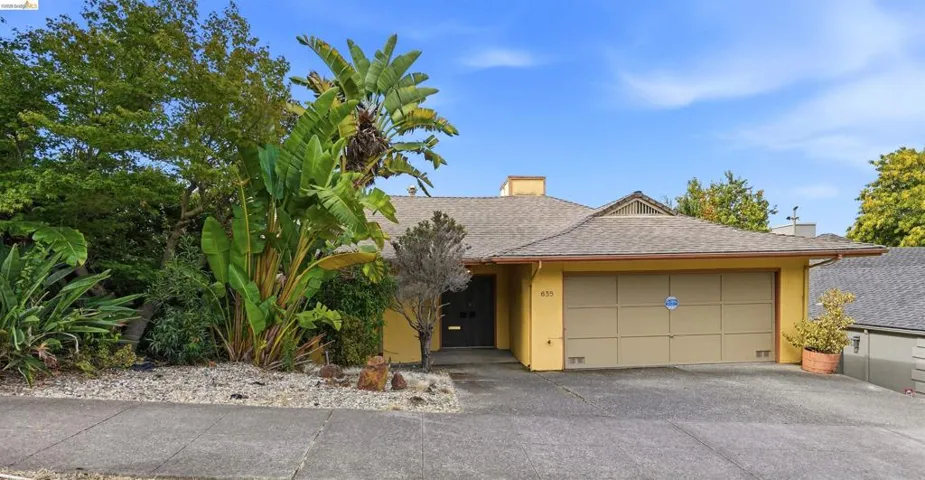 Single-story residence featuring a two-car garage, pitched shingle roof, and a mature landscaped front yard with varied plantings