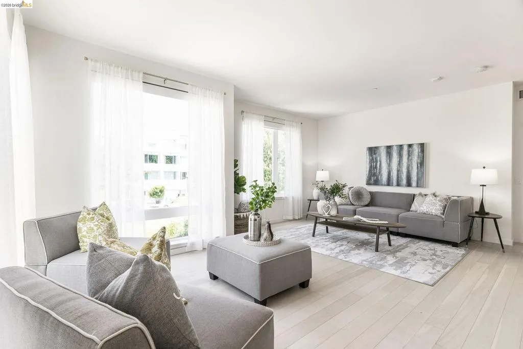 Spacious living area featuring light wood-finish flooring, white walls, and large windows with sheer white drapery