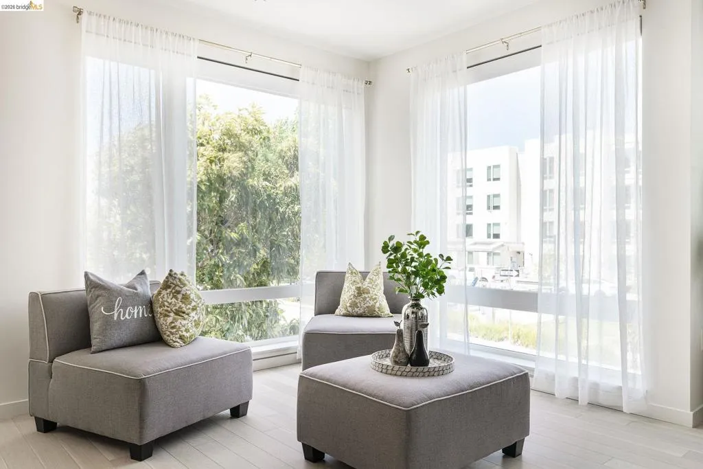 Bright living space featuring expansive windows with white sheer curtains, light wood-finish flooring, and crisp white walls
