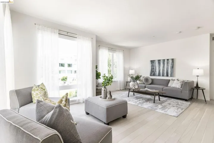 Spacious living area featuring light wood-finish flooring, white walls, and large windows with sheer white drapery