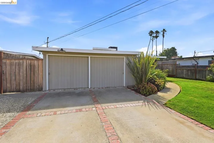 Concrete driveway with brick detailing, two-car garage with panel doors, mature landscaping, manicured lawn, and wood fencing