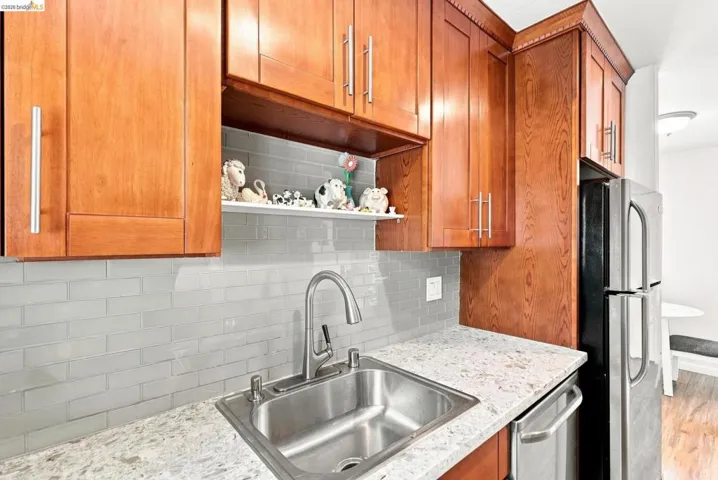 Compact kitchen featuring warm wood-finish cabinetry with chrome hardware, subway tile backsplash, light stone countertops, and stainless steel double-basin sink with pull-down faucet