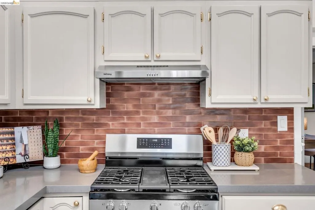 White paneled cabinetry with brass hardware, stainless steel range and hood, gray countertops, and a brick-patterned backsplash