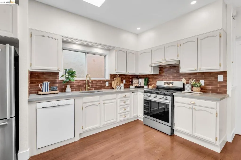 White cabinetry with brass-finish hardware, stainless steel appliances, brick-style backsplash, light-toned countertops, and wood-finish flooring