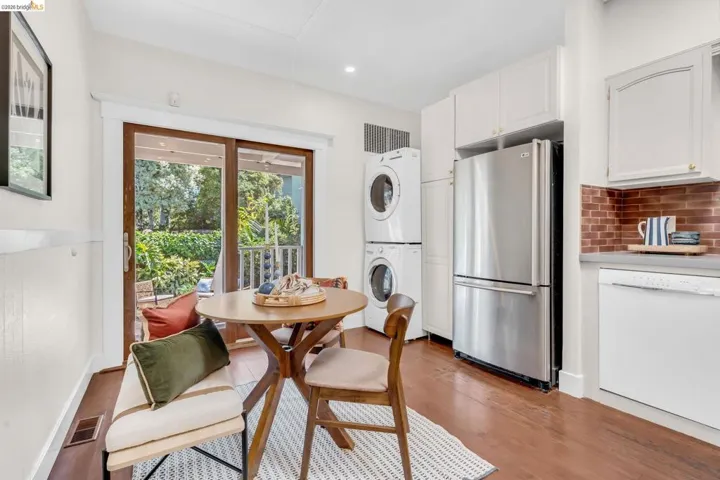 White shaker cabinetry with gold-tone hardware, stainless steel refrigerator, brick-finish backsplash, wood-finish flooring, and white stacked laundry appliances