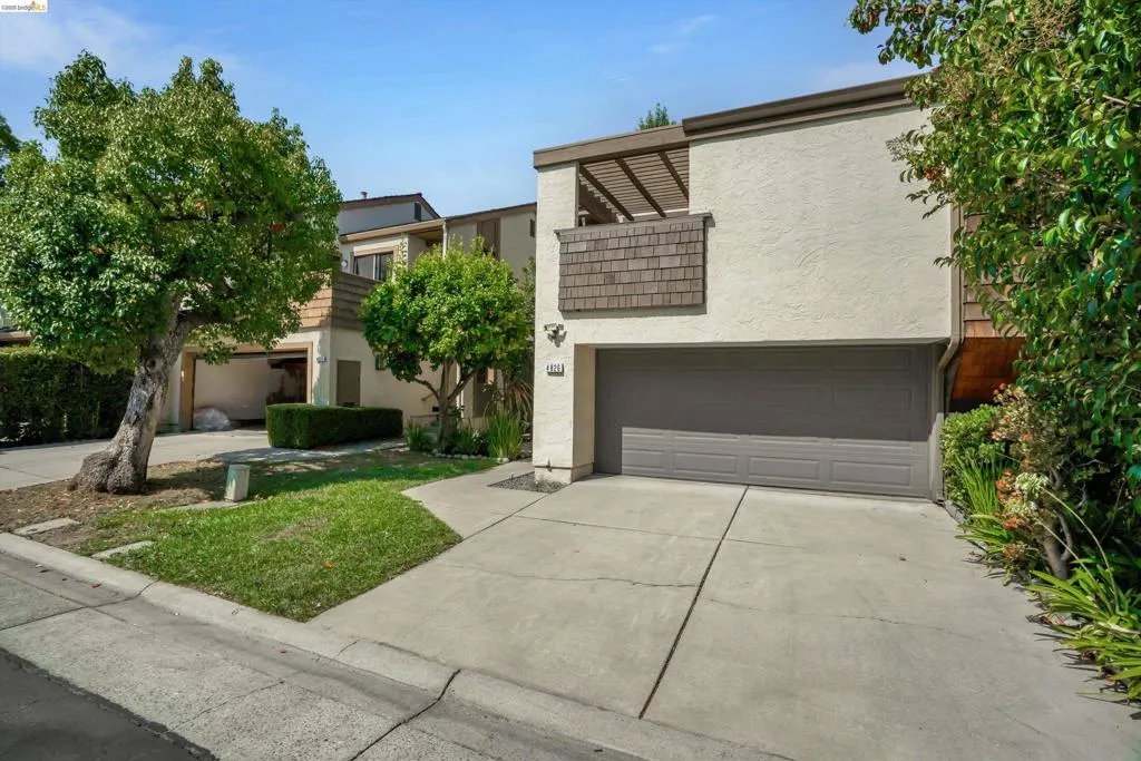 Stucco exterior with wood shingle accents, attached garage, concrete driveway, and mature landscaping
