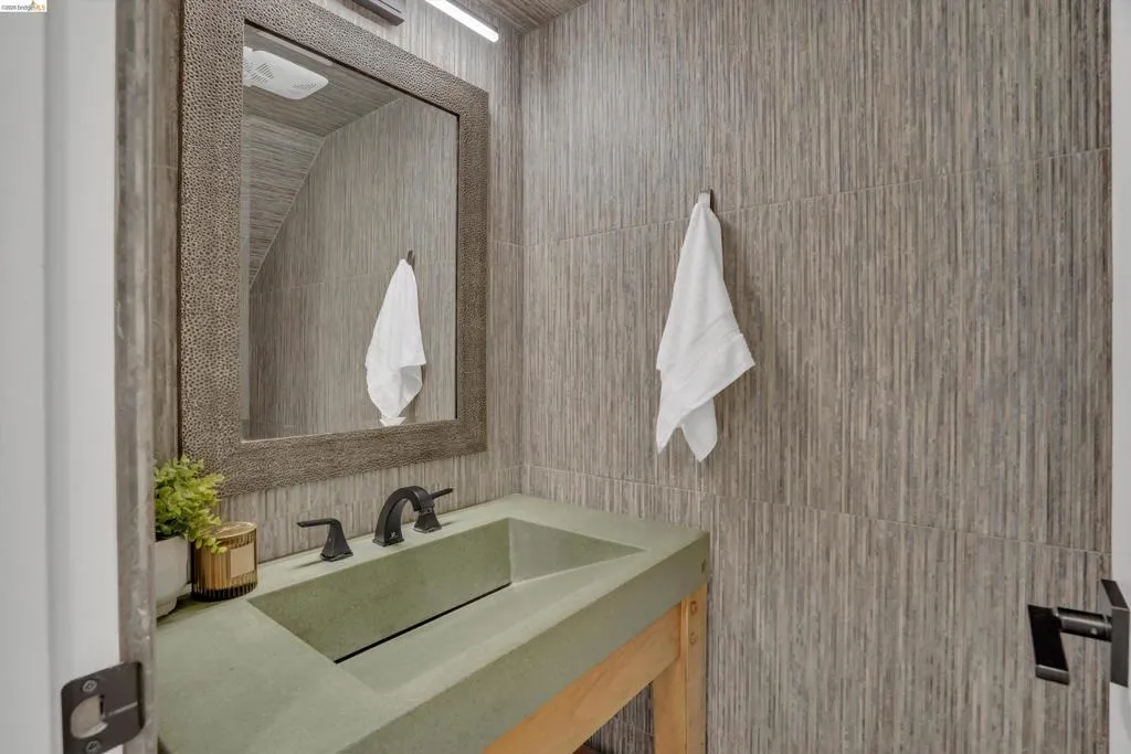 Textured wall tile, rectangular mirror with a decorative frame, integrated basin sink, black two-handle faucet, and black door hardware