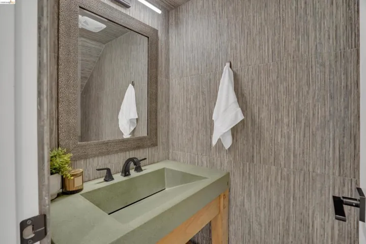 Textured wall tile, rectangular mirror with a decorative frame, integrated basin sink, black two-handle faucet, and black door hardware