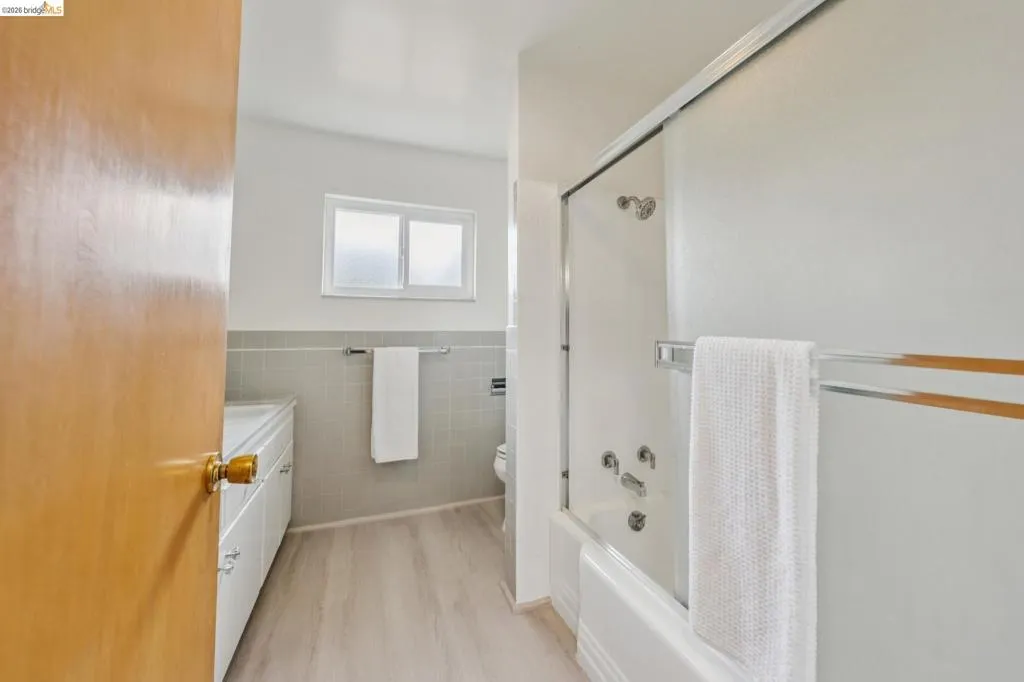 Bathroom featuring a white bathtub with a sliding glass door, light wood-finish flooring, white vanity with integrated sink, and a wall-mounted towel rack