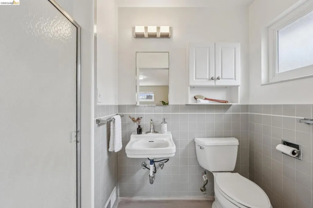 Shower enclosure with textured glass panel, wall-mounted sink, wall-mounted mirror, white wall cabinet, and light gray tiled walls