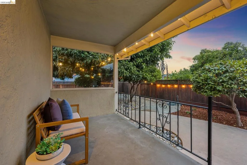 Covered patio with a concrete surface, decorative wrought iron railing, wood slat fencing, and mature trees