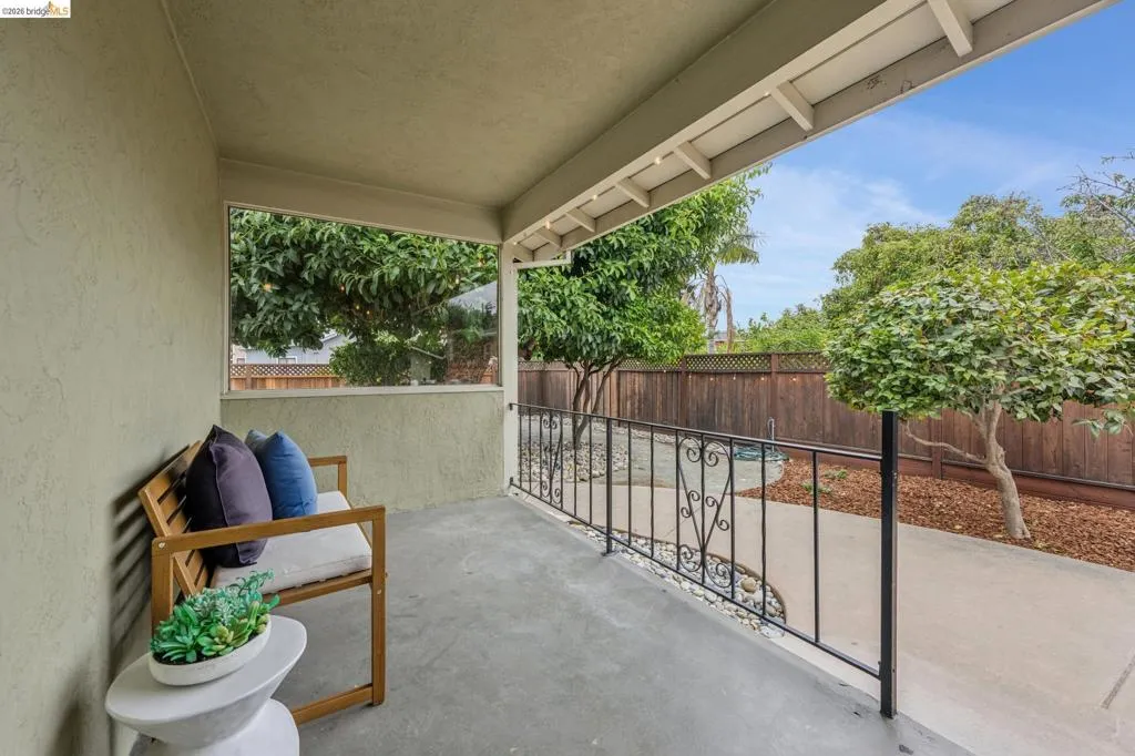 Covered patio area with concrete flooring, stucco wall finish, wrought iron railing, and mature landscaping