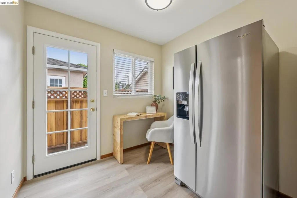 Light wood-finish flooring, a stainless steel side-by-side refrigerator, a white paneled door with glass inserts, a single window with blinds, and a built-in overhead light fixture