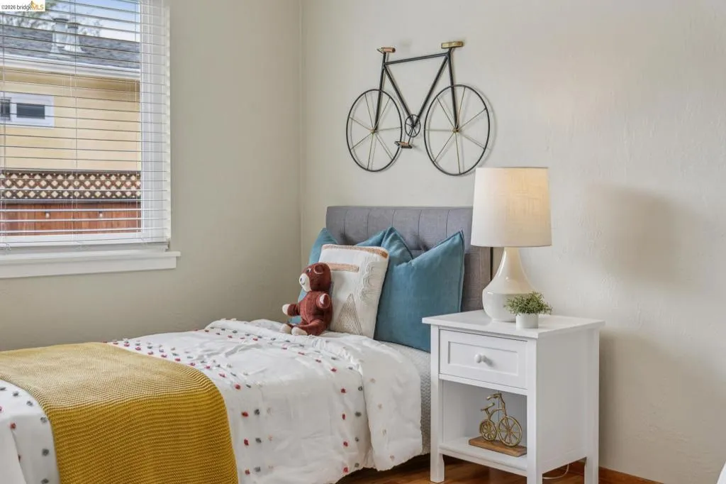 Window with horizontal blinds, light-toned wall finish, wood-finish flooring, and a gray upholstered headboard