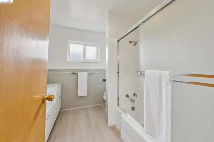 Bathroom featuring a white bathtub with a sliding glass door, light wood-finish flooring, white vanity with integrated sink, and a wall-mounted towel rack