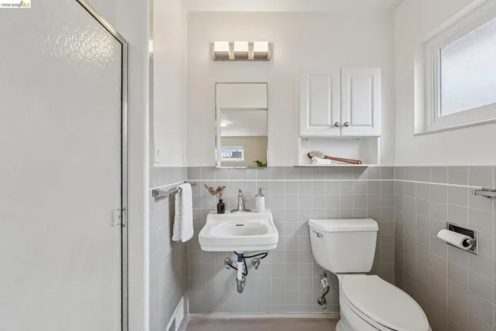 Shower enclosure with textured glass panel, wall-mounted sink, wall-mounted mirror, white wall cabinet, and light gray tiled walls