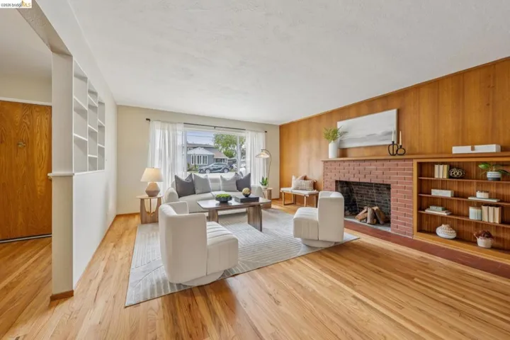 Hardwood flooring throughout, a red brick fireplace with a wood mantel, a large picture window, a partial wall with built-in shelving, and a wood-paneled accent wall