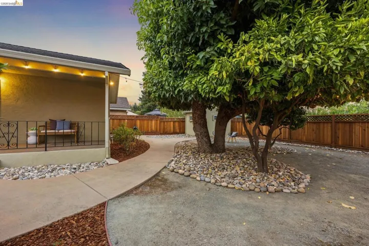 Covered patio with string lighting, wrought iron railing, concrete pathway, decorative rock landscaping, and wood privacy fencing