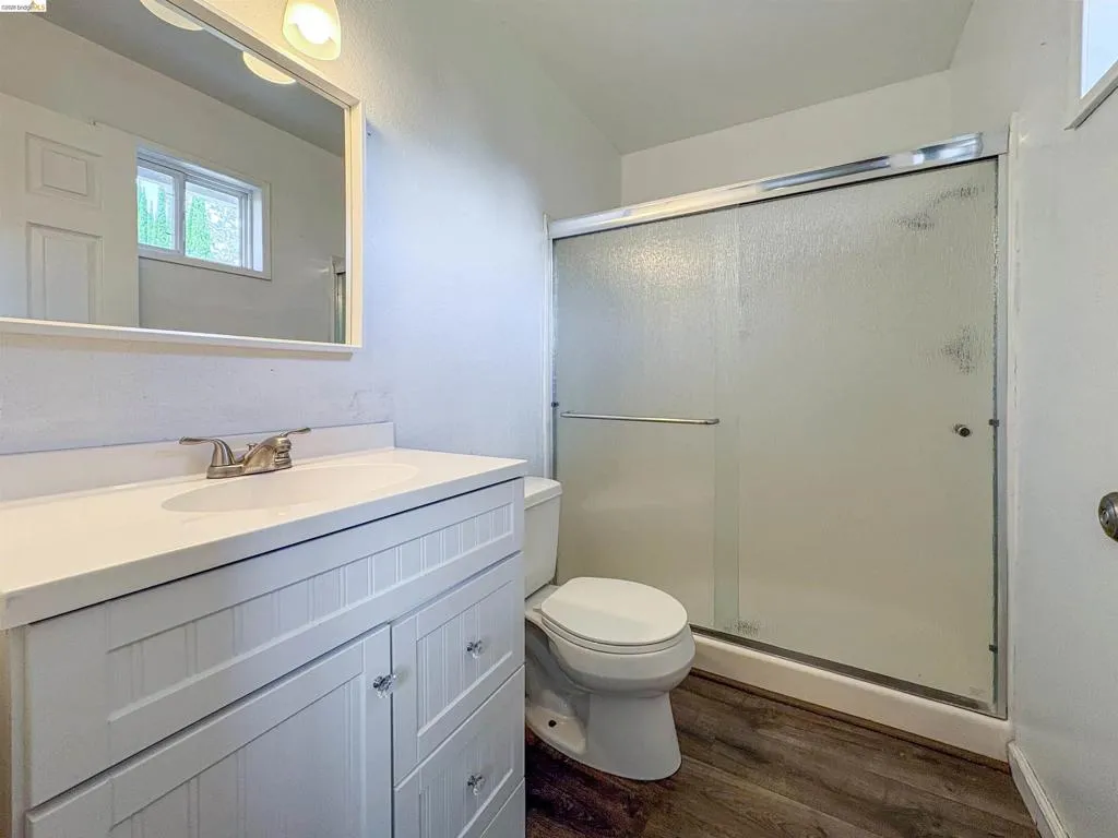 Bathroom featuring a white vanity with beadboard-style cabinetry, an integrated sink, a full-width mirror, a glass-enclosed shower, and wood-finish flooring
