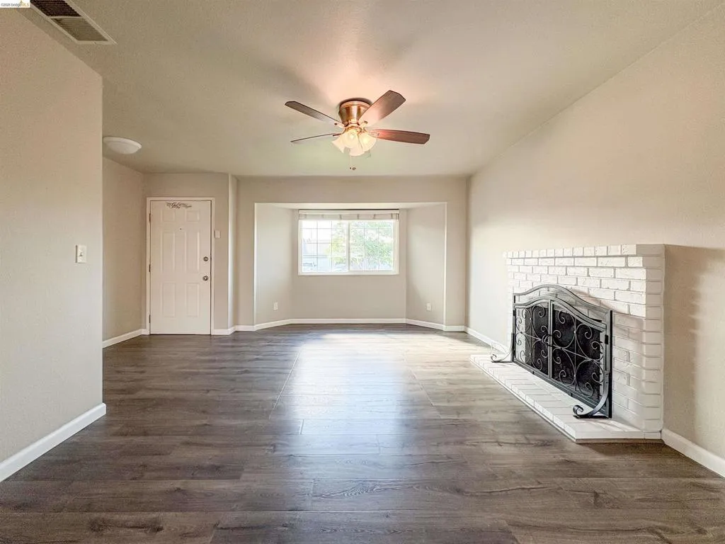 Spacious living area featuring wood-finish flooring, a white brick fireplace with a decorative screen, a ceiling fan with integrated lighting, and multiple windows