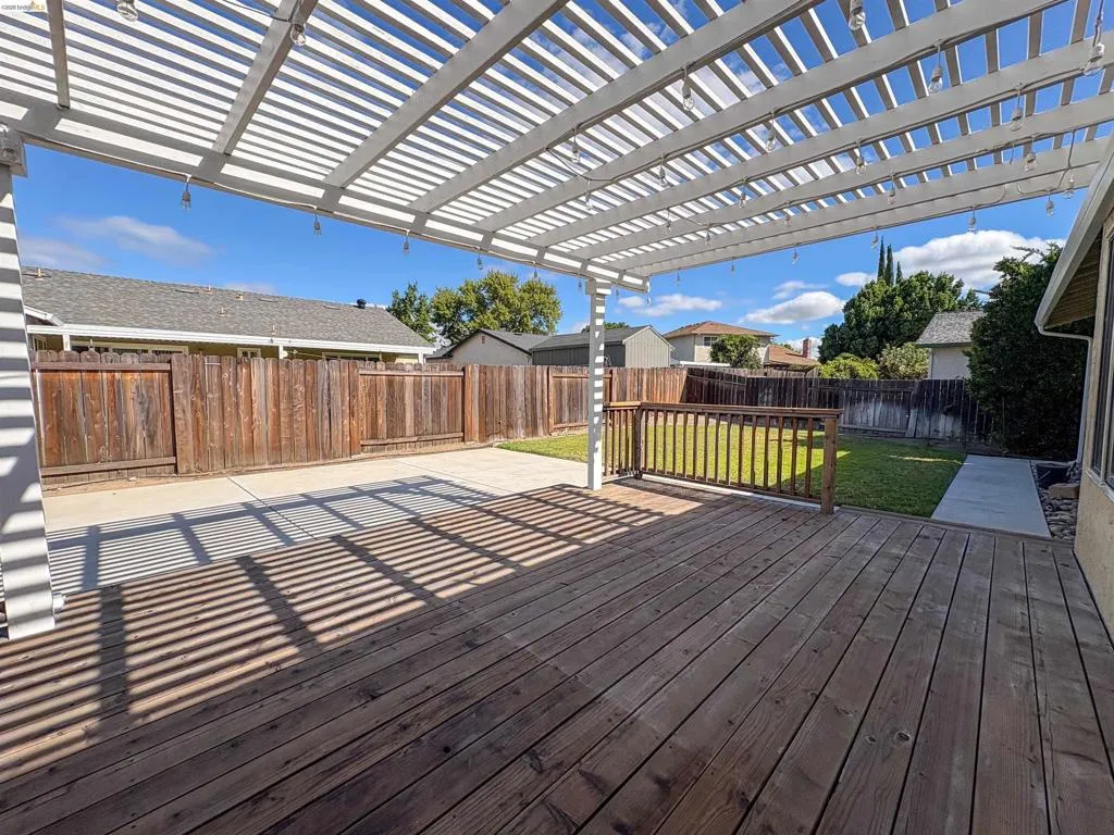 Exterior wood deck with a white pergola, concrete patio area, and a fenced backyard with a grass lawn
