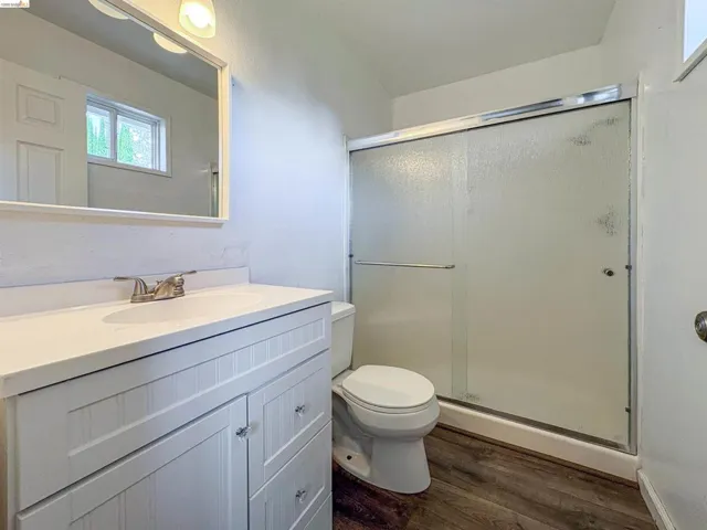 Bathroom featuring a white vanity with beadboard-style cabinetry, an integrated sink, a full-width mirror, a glass-enclosed shower, and wood-finish flooring