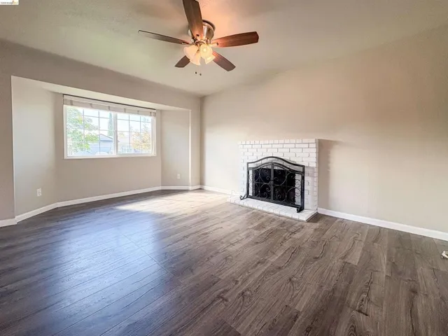 Wood-finish flooring, white brick fireplace, ceiling fan with integrated lighting, and a large window with white trim