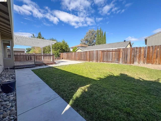 Expansive backyard featuring a wood deck, white pergola, concrete patio, and a manicured lawn