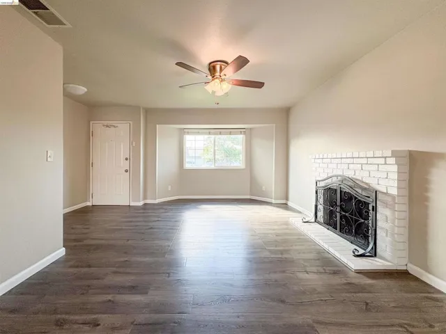 Spacious living area featuring wood-finish flooring, a white brick fireplace with a decorative screen, a ceiling fan with integrated lighting, and multiple windows