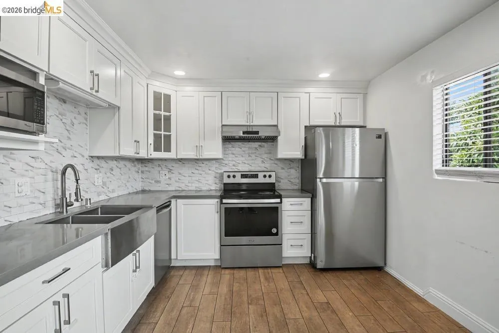 Kitchen with white cabinetry, wood-finish flooring, stainless steel appliances, a farmhouse sink, and a white tile backsplash