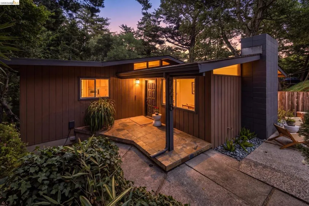 Mid-century modern exterior featuring wood siding, a stone-tiled entry, a prominent chimney with dark masonry, and abundant natural landscaping