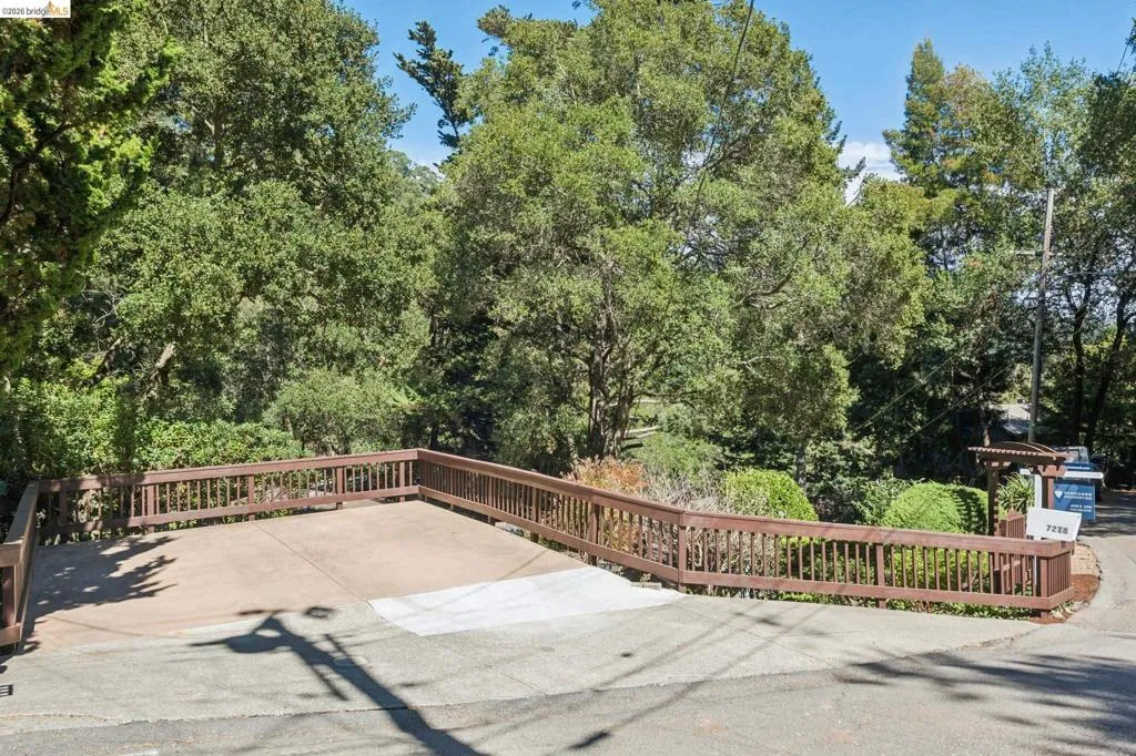 Paved driveway with a wood-stained railing, surrounded by mature trees and lush landscaping