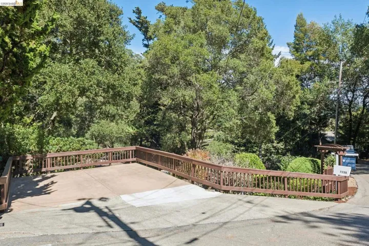 Paved driveway with a wood-stained railing, surrounded by mature trees and lush landscaping