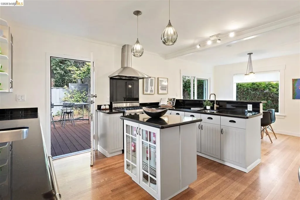 Hardwood flooring, stainless steel range hood, built-in kitchen island with glass-front cabinetry, black countertops, and direct access to an outdoor deck Hardwood flooring, stainless steel range hood, built-in kitchen island with glass-front cabinetry, black countertops, and direct access to an outdoor deck
