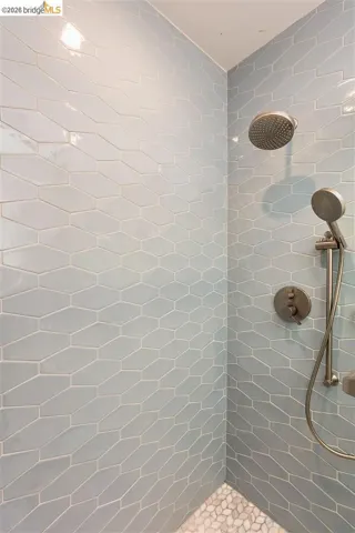 Shower with light blue elongated hexagonal wall tiles, pebble-style floor tiles, and a brushed nickel shower head with a handheld sprayer