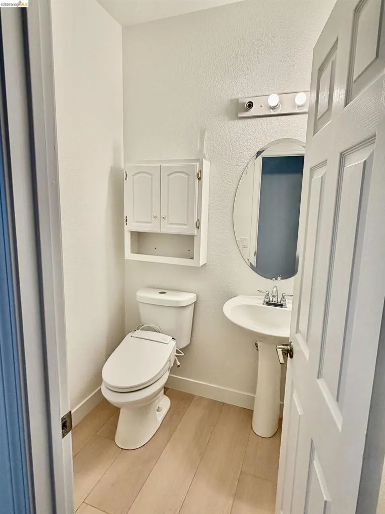 Pedestal sink with chrome faucet, oval mirror, wall-mounted light fixture, white wood-finish wall cabinet, and wood-finish flooring