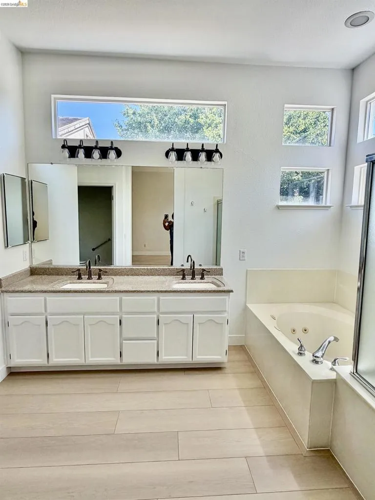 Dual vanity with granite-style countertop and undermount sinks, wood-finish flooring, built-in soaking tub, and multiple transom windows